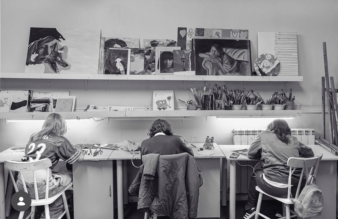 Tres alumnas dibujando de espaldas en la mesa larga del taller, con pinceles y trabajos en las baldas
